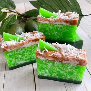 May include: Three bars of handmade soap with a lime and coconut scent. The soap is layered with green, brown, and white swirls and topped with a lime slice.