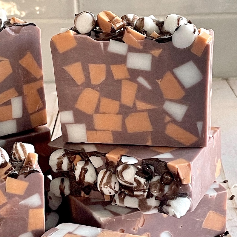 Puede incluir: Primer plano de varias barras de jab&oacute;n hechas a mano. El jab&oacute;n es de color marr&oacute;n oscuro con piezas blancas y naranjas en forma de mosaico. La parte superior de cada barra est&aacute; decorada con malvaviscos blancos, salsa de chocolate y trozos de caramelo.