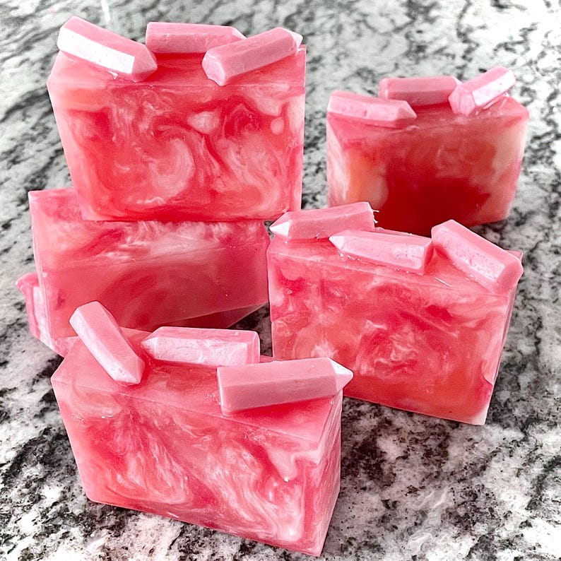 Pode incluir: V&aacute;rias barras de sab&atilde;o rosa e branco com redemoinhos, cada uma coberta com pequenos detalhes rosa em forma de cristal. Os sabonetes t&ecirc;m uma apar&ecirc;ncia marmorizada e s&atilde;o dispostos sobre uma superf&iacute;cie cinza salpicada. A est&eacute;tica geral &eacute; limpa e moderna.
