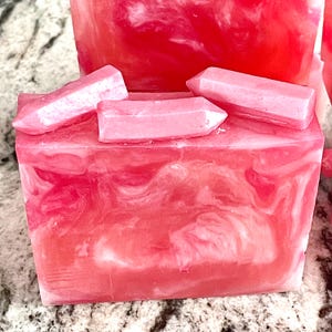 Pode incluir: Barra de sab&atilde;o rosa com um design em espiral e detalhes em forma de cristal rosa claro no topo. O sab&atilde;o tem uma apar&ecirc;ncia marmorizada com tons de rosa e branco. O sab&atilde;o tem formato de cubo.