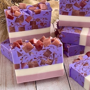 May include: Artisan soap bars in shades of purple, white, and pink, topped with reddish-brown mosaic-like pieces. The bars are stacked, showcasing the layered design. The soap has a textured surface and a decorative appearance.