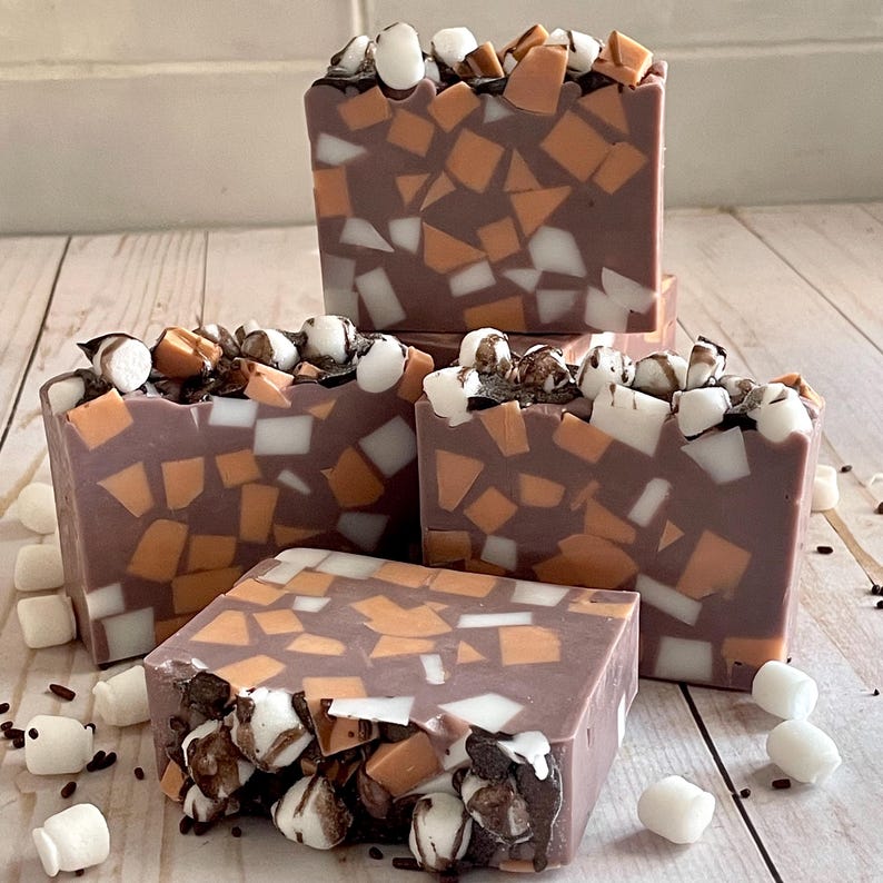 Puede incluir: Cuatro barras de jab&oacute;n cuadradas con un dise&ntilde;o de mosaico en tonos marr&oacute;n, naranja y blanco. El jab&oacute;n est&aacute; cubierto con malvaviscos blancos, chispas de chocolate y trozos de caramelo. Los jabones est&aacute;n colocados sobre una superficie de madera clara.
