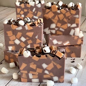 Puede incluir: Varias barras de jab&oacute;n hechas a mano con una base marr&oacute;n chocolate, incrustadas con piezas de mosaico naranja, blanco y marr&oacute;n. Cada barra est&aacute; cubierta con malvaviscos blancos y un chorrito de chocolate. Los jabones est&aacute;n colocados sobre una superficie de madera clara.