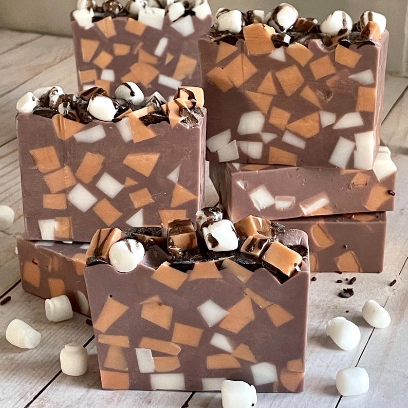 Puede incluir: Varias barras de jab&oacute;n hechas a mano con una base marr&oacute;n chocolate, incrustadas con piezas de mosaico naranja, blanco y marr&oacute;n. Cada barra est&aacute; cubierta con malvaviscos blancos y un chorrito de chocolate. Los jabones est&aacute;n colocados sobre una superficie de madera clara.