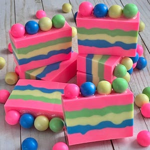 May include: A stack of colorful layered soap bars with a pink base, green and yellow stripes, and blue accents. Each bar is topped with a row of colorful candy-like balls.