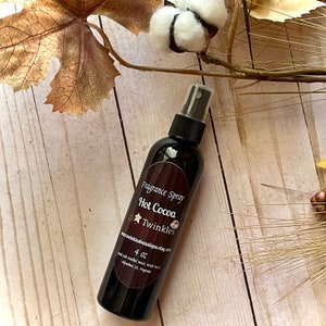 May include: A dark brown fragrance spray bottle with a label that reads "Fragrance Spray Hot Cocoa Twinkles". The bottle contains 4 oz of product. The background includes fall foliage and a wooden surface.