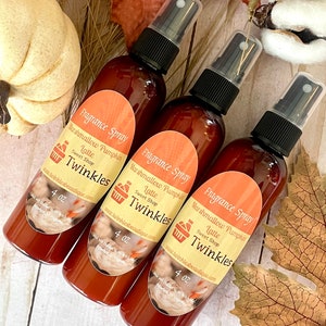 May include: Three 4 ounce bottles of brown liquid fragrance spray with white labels. The labels feature a brown and white illustration of a cup of coffee with whipped cream and a shopping cart icon. The text on the labels reads "Fragrance Spray", "Marshmallow Pumpkin Latte", "Sweet Shop", and "Twinkles". The bottles are on a light brown wooden surface.