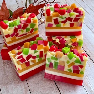 May include: Several bars of handmade soap with a colorful mosaic design. The soap features layers of red, white, and yellow, topped with a mix of red, green, orange, and pink soap pieces. The bars are stacked on a light wood surface.