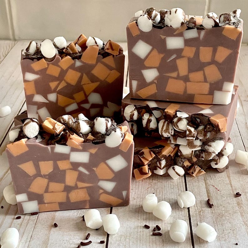 Puede incluir: Cuatro barras de jab&oacute;n artesanal con tem&aacute;tica de chocolate. El jab&oacute;n es de color marr&oacute;n oscuro con trozos naranjas, blancos y marrones incrustados. La parte superior de cada barra est&aacute; decorada con malvaviscos blancos y un chorrito de chocolate.