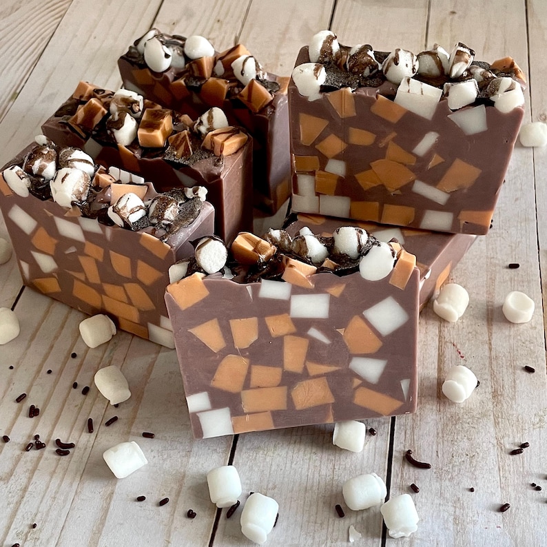 Puede incluir: Cinco barras de jab&oacute;n artesanal con tem&aacute;tica de chocolate. El jab&oacute;n es marr&oacute;n con piezas de mosaico naranja y blanco. Coberturas: malvaviscos, trozos de caramelo y salsa de chocolate. Malvaviscos y chispas de chocolate esparcidos sobre una superficie de madera.