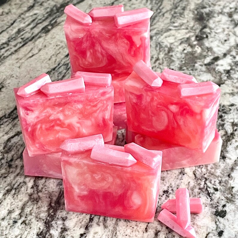 Pode incluir: V&aacute;rias barras de sab&atilde;o rosa e branco com redemoinhos s&atilde;o empilhadas, cada uma coberta com enfeites em forma de cristal rosa claro. O sab&atilde;o tem uma apar&ecirc;ncia marmorizada, com diferentes tons de rosa. Alguns cristais est&atilde;o espalhados ao redor da base da pilha.