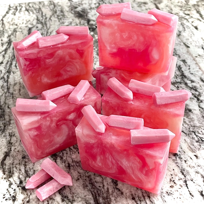 Pode incluir: V&aacute;rias barras de sab&atilde;o rosa e branco com redemoinhos, cada uma coberta com m&uacute;ltiplas decora&ccedil;&otilde;es rosa em forma de cristal. Os sabonetes s&atilde;o dispostos em uma pilha, com algumas barras espalhadas. O fundo &eacute; uma superf&iacute;cie cinza e branca salpicada.