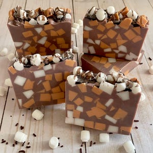 Puede incluir: Cuatro barras cuadradas de jab&oacute;n artesanal con base marr&oacute;n chocolate, incrustadas con trozos naranjas, blancos y marrones. La parte superior de cada barra est&aacute; decorada con malvaviscos blancos y un chorrito de chocolate. Malvaviscos y chispas esparcidos.