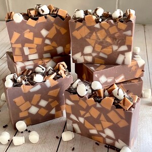 Puede incluir: Barras de jab&oacute;n artesanal con base marr&oacute;n chocolate, incrustadas con piezas de mosaico blancas, naranjas y marrones. Cubiertas con malvaviscos, salsa de chocolate y cuadrados de chocolate. Se ven seis barras apiladas.