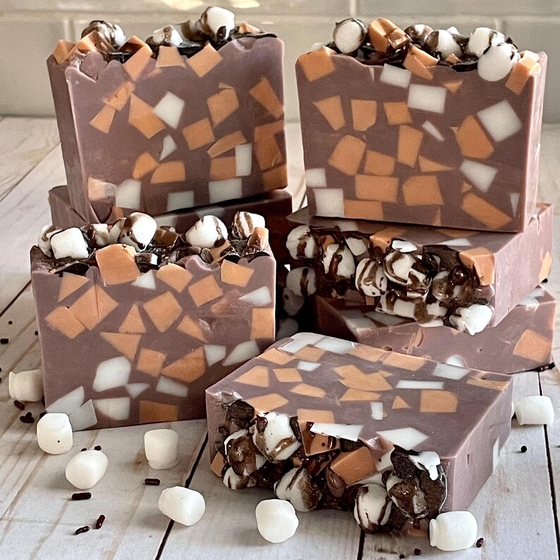 Puede incluir: Varias barras de jab&oacute;n hechas a mano con tem&aacute;tica de chocolate y malvavisco. Las barras son de color marr&oacute;n oscuro, con piezas de mosaico naranja y blanco incrustadas. Las tapas est&aacute;n decoradas con malvaviscos y un chorrito de chocolate. Malvaviscos esparcidos por la base.