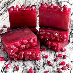 May include: A stack of four red soap bars with a glittery, crystal-like design on top. The soap bars are on a white marble surface with small red crystals scattered around.