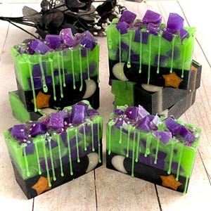 May include: Rectangular bars of handmade soap featuring a black base with a crescent moon and star design, topped with a vibrant green layer, purple translucent chunks, and green drips. The soap is decorated with small star-shaped glitter pieces.