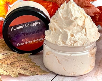 May include: A glass jar filled with a creamy white shea body butter. The jar has a black lid with a label that reads "Autumn Campfire" and "Twinkles Shea Body Butter".