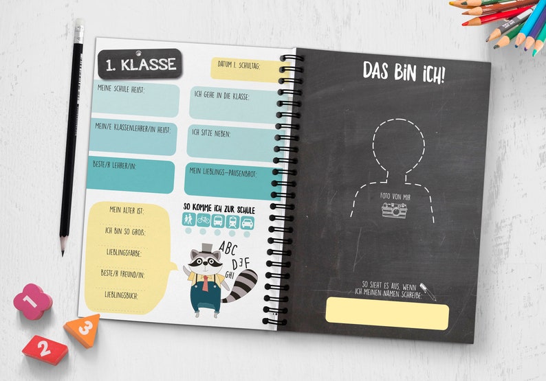 Op de afbeelding: Een zwart-wit schoolschrift met een gele en blauwe omslag. De omslag heeft de tekst "1. Klasse" en "Das bin ich!" Het schrift heeft een spiraalbinding en een wasbeerillustratie.