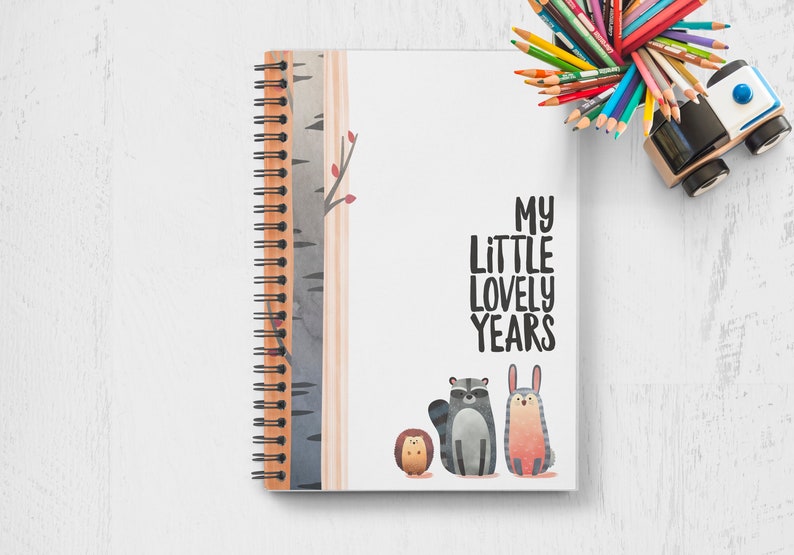 Puede incluir: Un cuaderno blanco de espiral con un tema de bosque. La portada presenta un abedul, un mapache, un erizo y un conejo. El texto "My Little Lovely Years" est&aacute; impreso en negro en la portada.