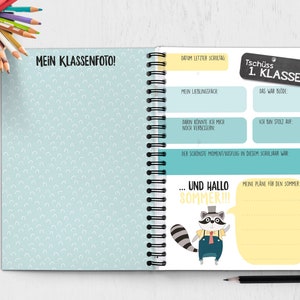 Op de afbeelding: Een blauw en geel notitieboek met een wasbeerillustratie en de tekst "Tsch&uuml;ss 1. Klasse" op de omslag. Het notitieboek heeft een spiraalbinding en is versierd met een lichtblauwe achtergrond met witte halve cirkels.