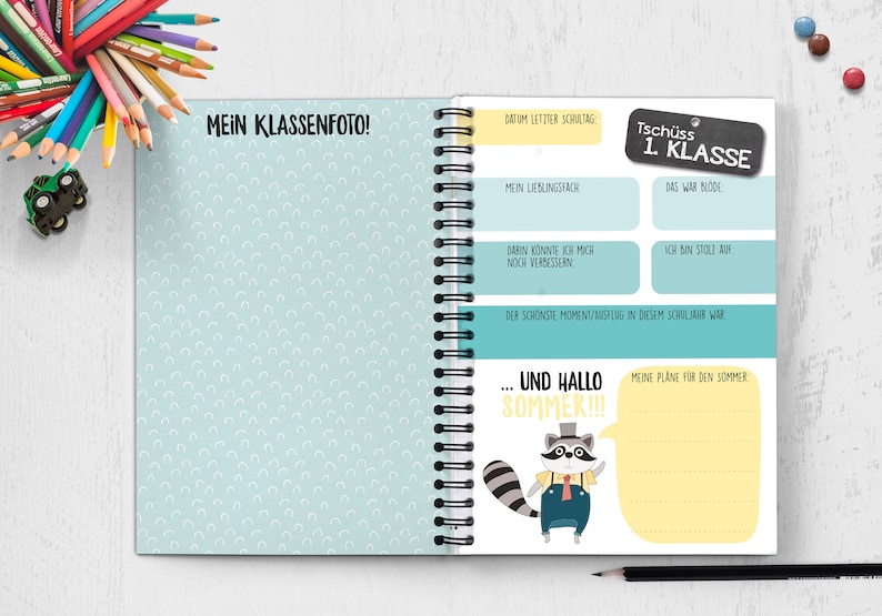 Op de afbeelding: Een blauw en geel notitieboek met een wasbeerillustratie en de tekst "Tsch&uuml;ss 1. Klasse" op de omslag. Het notitieboek heeft een spiraalbinding en is versierd met een lichtblauwe achtergrond met witte halve cirkels.