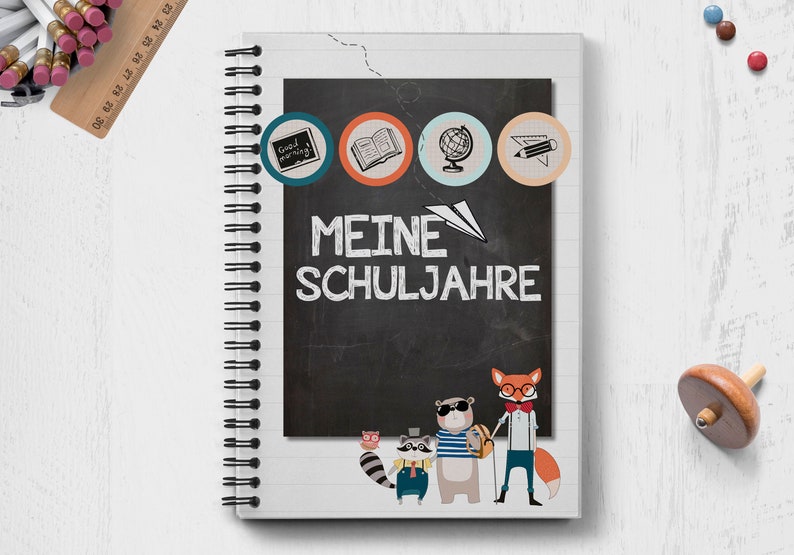 Op de afbeelding: Een wit spiraalnotitieblok met een zwarte schoolbordomslag. Het schoolbord heeft de tekst "MEINE SCHULJAHRE" erop geschreven. Bovenaan het schoolbord staan vier kleurrijke cirkels met pictogrammen die een boek, een wereldbol, een potlood en een klok voorstellen. Onderaan het schoolbord staan vijf cartoondieren.