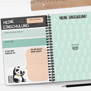 Op de afbeelding: Een groen en zwart spiraalnotitieblok met de tekst "Meine Einschulung!" op de omslag. De omslag heeft een patroon van witte wortels op een lichtgroene achtergrond. De binnenkant van het notitieblok heeft een zwarte schoolbordachtergrond met de tekst "Meine Einschulung" en een pandabeerillustratie.