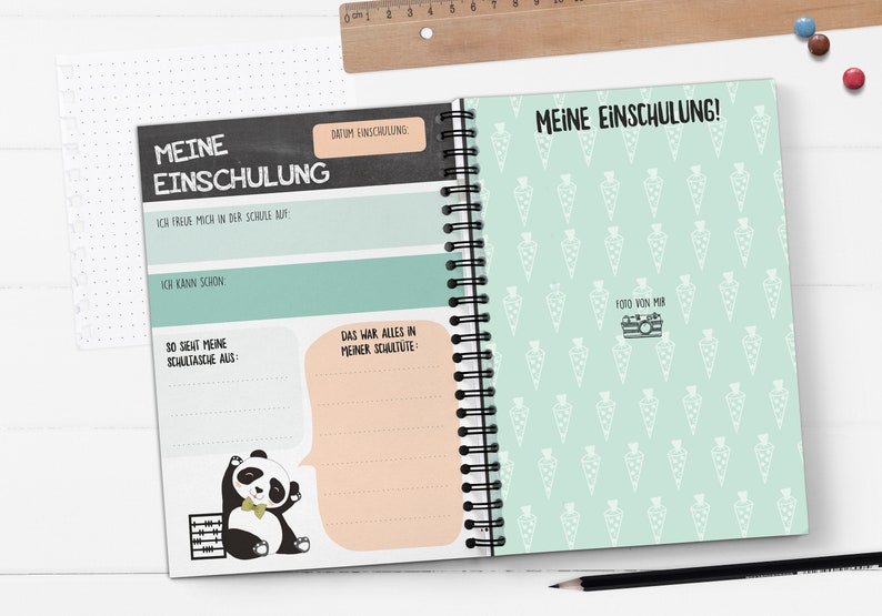 Op de afbeelding: Een groen en zwart spiraalnotitieblok met de tekst "Meine Einschulung!" op de omslag. De omslag heeft een patroon van witte wortels op een lichtgroene achtergrond. De binnenkant van het notitieblok heeft een zwarte schoolbordachtergrond met de tekst "Meine Einschulung" en een pandabeerillustratie.