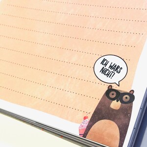 Puede incluir: Una libreta de notas marr&oacute;n claro con un patr&oacute;n de l&iacute;neas punteadas y un oso de dibujos animados con un globo de di&aacute;logo que dice "Ich wars nicht!" (&iexcl;No fui yo!).