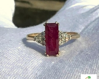 May include: A gold ring with a large, rectangular, deep red ruby as the central gemstone. Small, clear gemstones are set on either side of the ruby. The ring is displayed on a white surface.