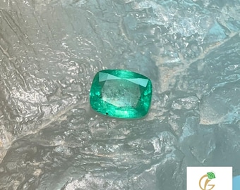 May include: A square-cut, vivid green emerald gemstone. The gem is set against a textured, light grey surface. The image includes the logo of "izna Gems" at the bottom right.