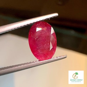 May include: A vibrant red oval-shaped gemstone held in a pair of tweezers. The gemstone has a faceted cut and a glossy finish.