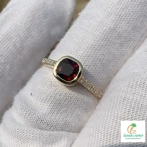 Könnte beinhalten: Ein Ring mit einem dunkelroten Edelstein im Kissenschliff, gefasst in eine polierte Goldfassung, mit einer Ringschiene, die an den Seiten mit kleinen Pavé-Steinen besetzt ist. Der Ring liegt auf einem weißen Stoffhintergrund mit einem Logo.