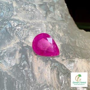 Puede incluir: Una gema de rubí facetada en forma de pera, de un vibrante color magenta. La gema se encuentra sobre una superficie texturizada de color gris azulado claro. La imagen incluye un logotipo en la esquina inferior derecha.