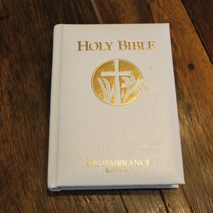 May include: White Holy Bible with gold lettering and a gold cross and lily design. The book says "Remembrance Edition" on the bottom.
