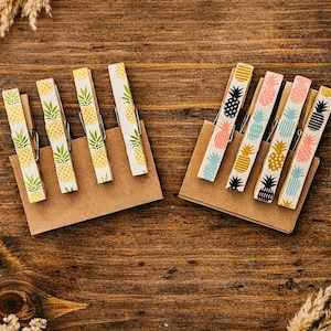 May include: Wooden clothespins with pineapple designs in yellow, black, and pink, arranged on brown cardstock. The clothespins are set against a dark wood background, creating a tropical theme.