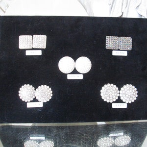 May include: A black velvet display board with six pairs of rhinestone embellishments. The embellishments are in various shapes and sizes, including round, square, and oval. The board has a white label with the text "2227" on it.