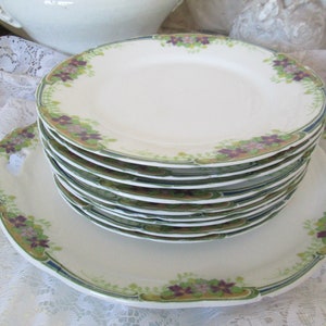 May include: A stack of vintage white porcelain plates with a floral design in shades of green, purple, and gold. The plates have a scalloped edge and a raised rim.