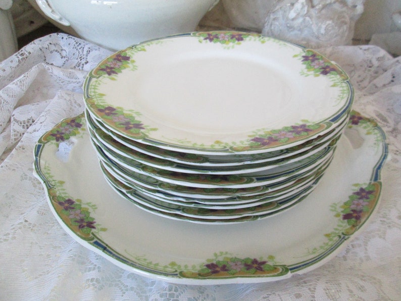 May include: A stack of vintage white porcelain plates with a floral design in shades of green, purple, and gold. The plates have a scalloped edge and a raised rim.
