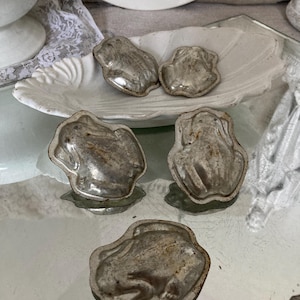 May include: Five vintage metal frog-shaped molds for making chocolate or candy. The molds are silver in color and have a worn, antique look.