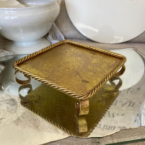 Peut inclure: Un petit plateau carré en métal doré avec une bordure en forme de corde. Le plateau a trois pieds et est posé sur une surface miroir.