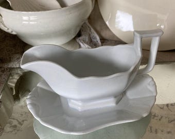 May include: A white porcelain gravy boat with a matching scalloped-edge saucer. The gravy boat has a handle and a pouring spout. Other white ceramic serving dishes are in the background.