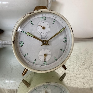 Op de afbeelding: Vintage crèmekleurige wekker met goudkleurige accenten en een verweerde afwerking. De wijzerplaat heeft zwarte cijfers, groene markeringen en de merknaam "Mantha Famos". De klok heeft een kleine secondenwijzer en staat op drie goudkleurige poten.