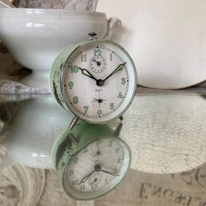 Könnte beinhalten: Ein grüner Vintage-Regina-Wecker mit weißem Zifferblatt, schwarzen Zeigern und grünen Ziffern. Die Uhr hat ein abgenutztes Finish und spiegelt sich in einer spiegelnden Oberfläche wider. Die Uhr ist ein klassisches Design.
