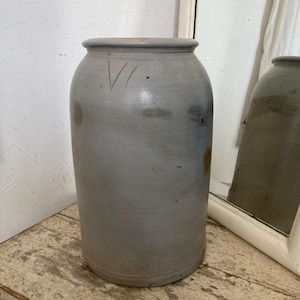 May include: A large, gray stoneware crock with a slightly textured surface. The crock has a wide mouth and a rounded bottom. There are faint markings on the side of the crock.