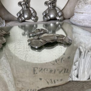 May include: Three silver teddy bear figurines on a mirrored surface. The bears are sitting up and have a classic teddy bear design.