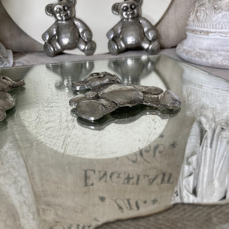 May include: Three silver teddy bear figurines on a mirrored surface. The bears are sitting up and have a classic teddy bear design.