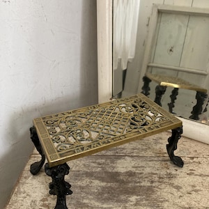 May include: A brass trivet with an ornate openwork design and black cast iron legs. The trivet has the words "HAMES" and "BEST" around the edges. The legs are decorative and have a floral design.