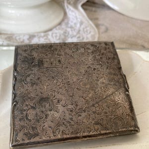 Könnte beinhalten: Ein silbernes Zigarettenetui mit einem gravierten Blumenmuster und den Initialen "D.K." auf der Vorderseite.